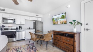 Een compacte keuken met witte kastjes, een magnetron, een kookplaat en een koelkast. Er is een eethoek met een ronde tafel en rieten stoelen. Een houten kast met lades staat tegen de muur, en er staat een bloempot op de bovenkant.