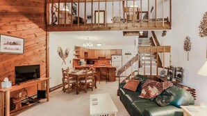 A spacious living area with a green leather sofa, wooden dining table, and a TV on a wooden stand. A kitchen with wooden cabinets and a staircase leading to an upper level is visible. - Cozy Mountain Condo w/Outdoor Heated Pool Access (Fraser)
