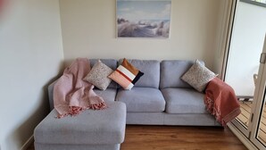 A grey sectional sofa with a throw blanket and patterned pillows, a framed landscape painting on the wall, and a wooden floor. - Beaton's Beach Front Studio - Inverloch (Inverloch)