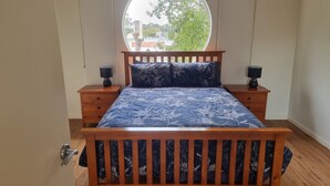 A bedroom with a wooden bed, two nightstands, a round window, and a view of greenery outside. - Beaton's Beach Front Studio - Inverloch (Inverloch)