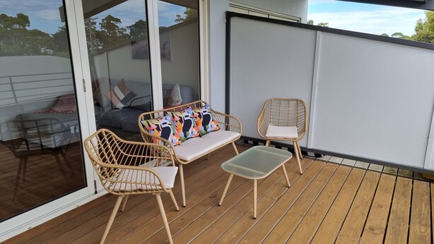 An outdoor seating area with wicker chairs and a cushioned bench, a glass door leading to a covered patio, and a view of the sky and trees. - Beaton's Beach Front Studio - Inverloch (Inverloch)