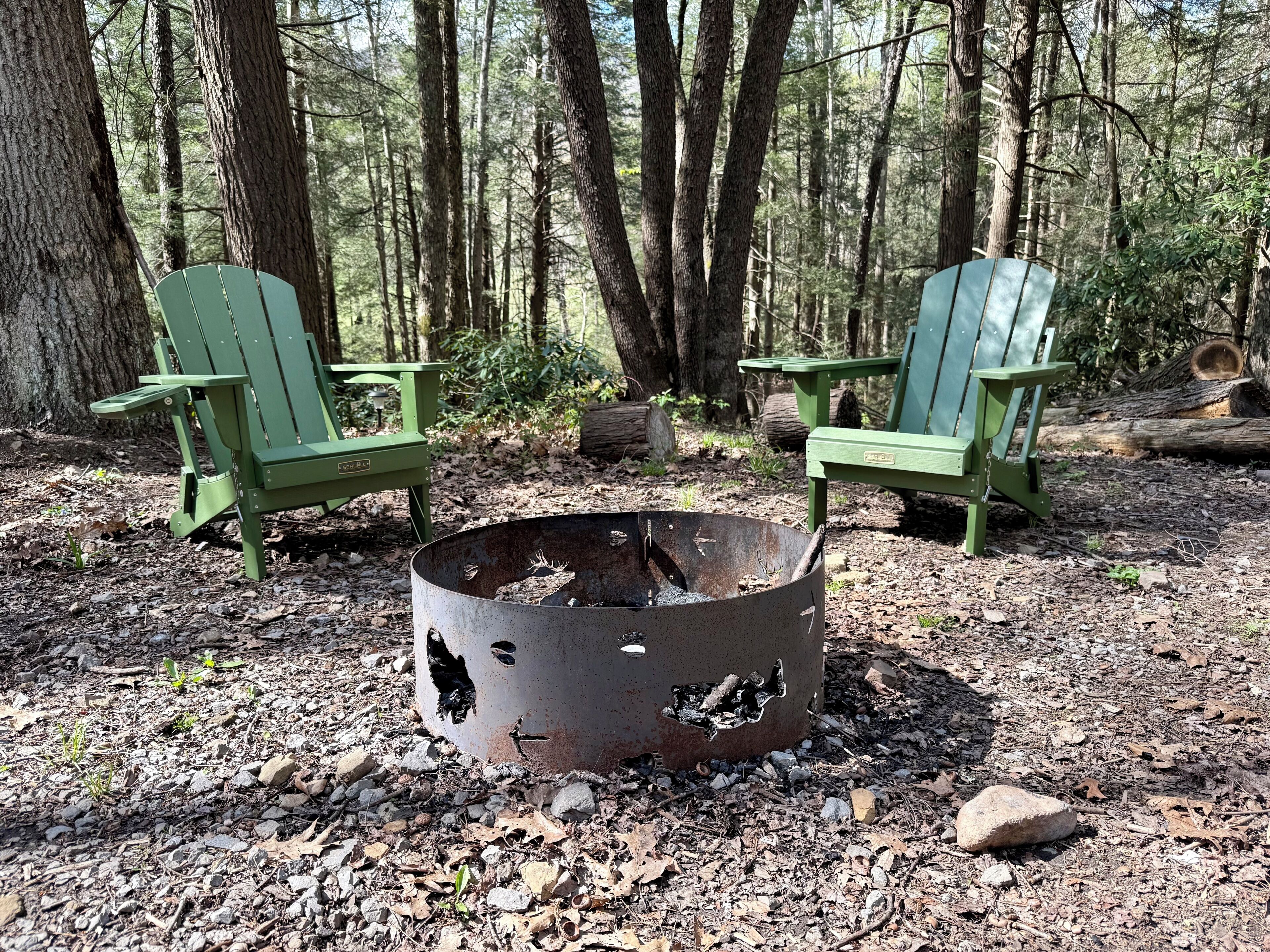 Dos sillas Adirondack verdes, un brasero de metal y un área boscosa.