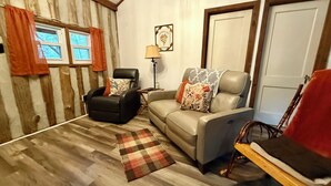 Un salon chaleureux avec un fauteuil en cuir noir, un canapé beige et un fauteuil à bascule en bois. Un tapis à carreaux recouvre le sol, et les murs sont recouverts de panneaux en bois.