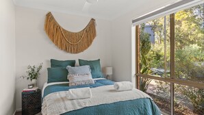 A bedroom with a large window, a bed with teal bedding, a woven wall hanging, and a bedside plant. - Coastal Sounds at Mollymook (Mollymook Beach)
