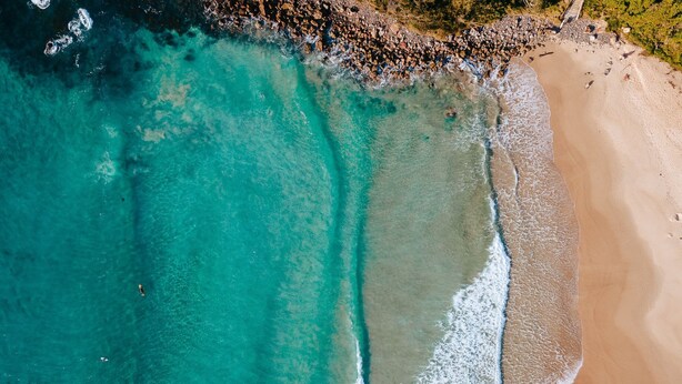 An aerial view of a beach with clear turquoise waters, a sandy shore, and a rocky area with people. - Coastal Sounds at Mollymook (Mollymook Beach)
