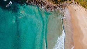 An aerial view of a beach with clear turquoise waters, a sandy shore, and a rocky area with people.