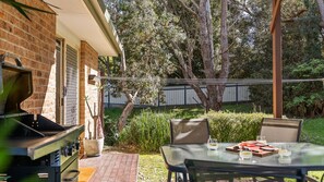 An outdoor patio with a grill, a table with chairs, and a view of a backyard with trees and a fence.