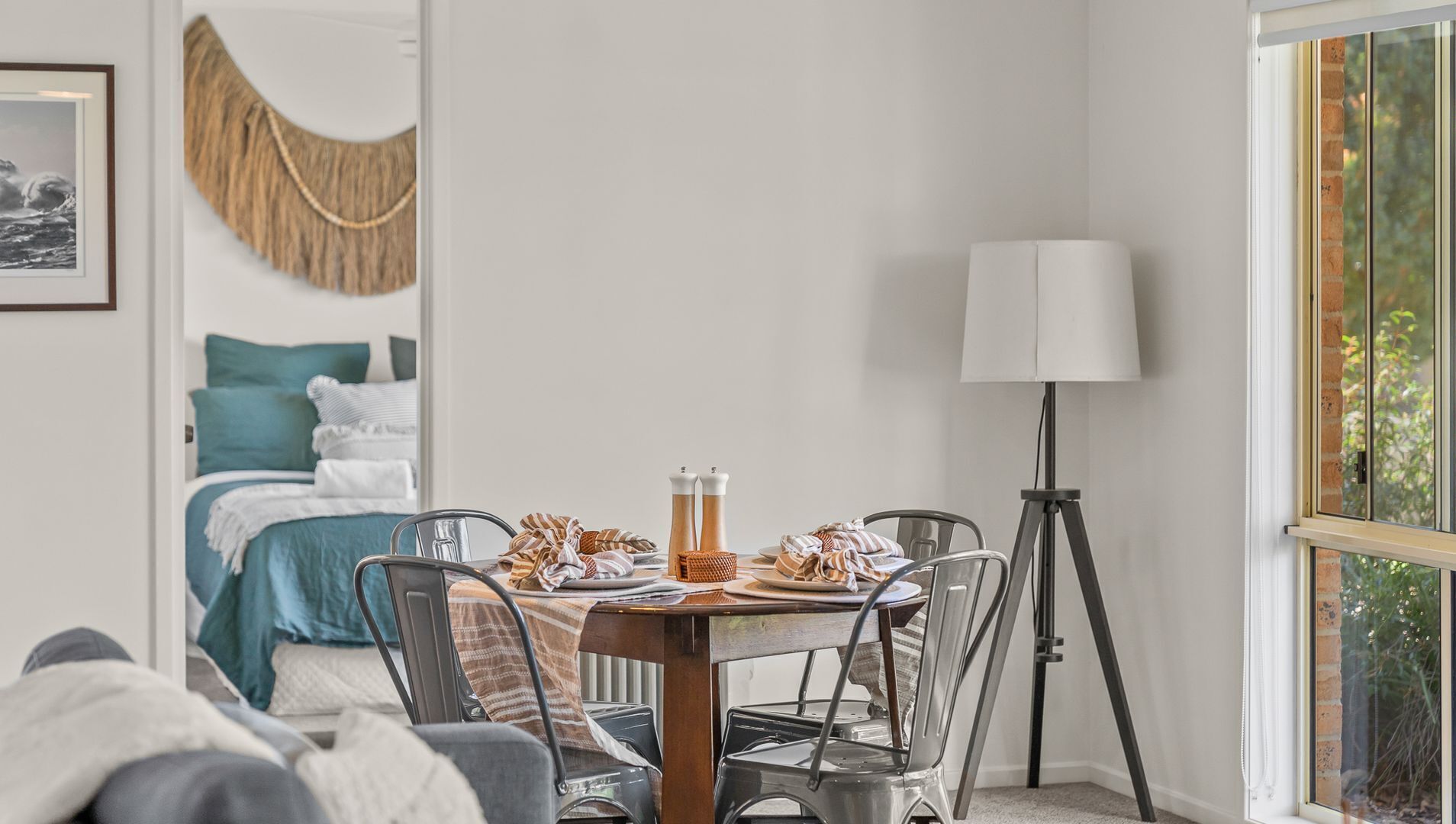 A neatly arranged dining area with a table set for two, a bed with blue bedding, a lamp, and a view of greenery outside.
