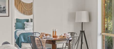 A neatly arranged dining area with a table set for two, a bed with blue bedding, a lamp, and a view of greenery outside.