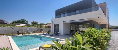 A modern two-story house with a swimming pool, outdoor seating, and a view of the surrounding landscape.