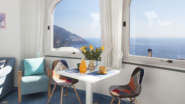 A room with a table and chairs, a vase with flowers, and a view of the sea through large windows. - Estate4home - Villa Settemari (Positano)