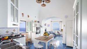A bright kitchen with a wooden table and white chairs, a sink, and a view of the sea through an arched window. - Estate4home - Villa Settemari (Positano)