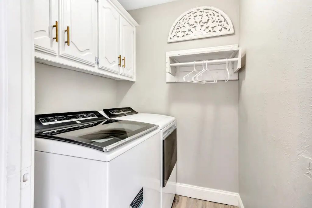 A laundry room with a white washing machine and dryer, white cabinets with gold handles, and a wall-mounted drying rack.