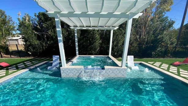 A swimming pool with a white pergola structure, surrounded by a tiled deck and a wooden fence.