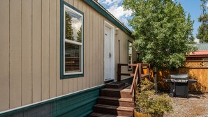 Une maison en bois avec une porte d’entrée, un escalier en bois menant à la porte, et un barbecue dans le jardin.