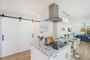 A modern kitchen with a central island, a stainless steel range hood, and a dining area with a blue sofa and a TV.