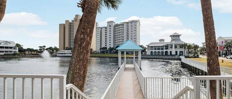 A wooden boardwalk leading to a gazebo over water, surrounded by palm trees and buildings.