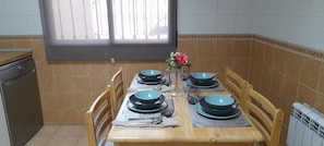A dining table set with plates, bowls, and cutlery, flanked by chairs, in a kitchen with tiled walls and a window.