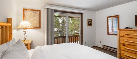 A bedroom with a bed, a wooden dresser, a nightstand with a lamp, a framed map on the wall, and a sliding glass door to the outside.