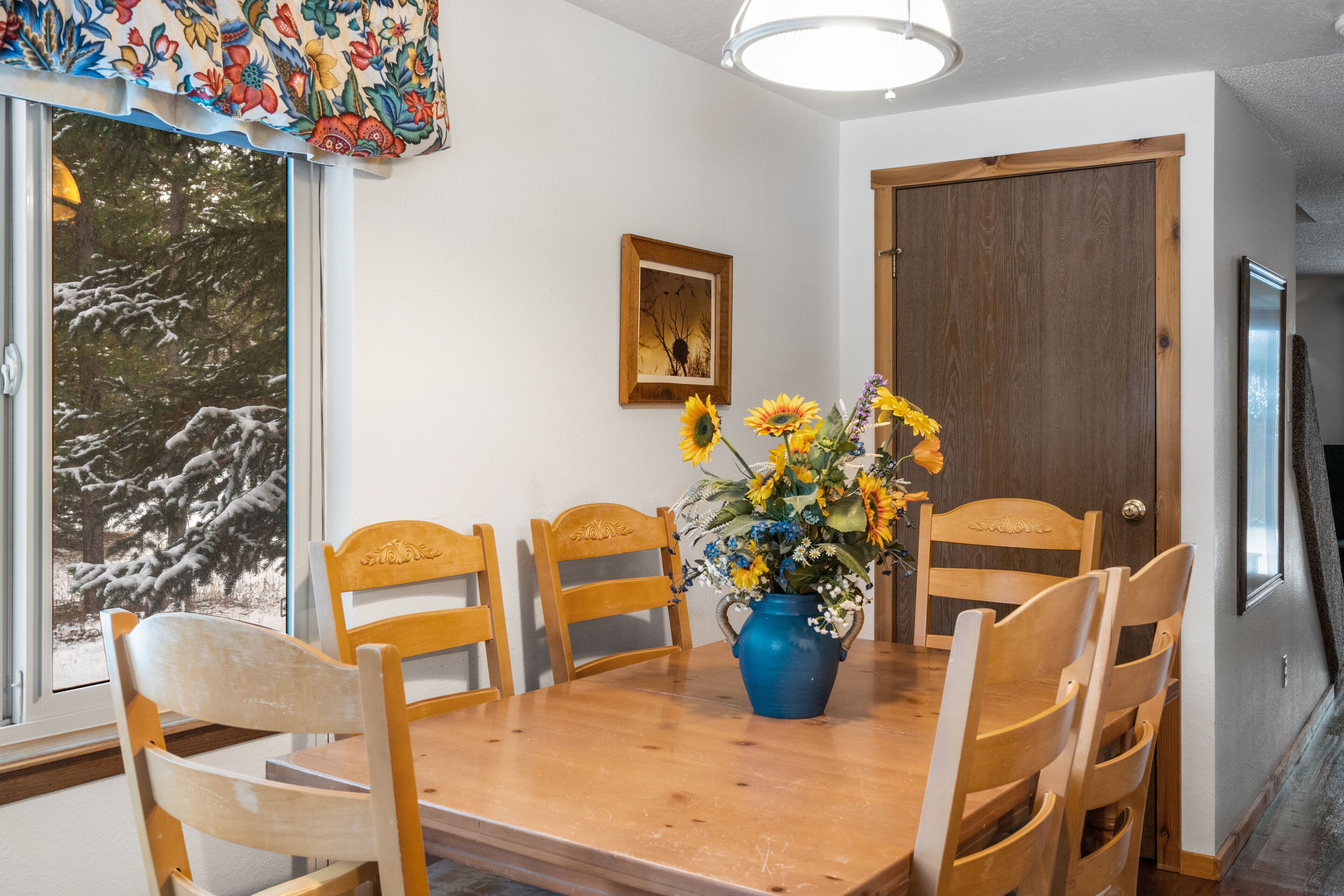 A dining area with a wooden table and chairs, a vase with flowers, a framed picture on the wall, and a window with a view of snow-covered trees.
