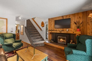 Una sala de estar con chimenea, sillas estampadas en verde, una escalera de madera y un televisor de pantalla plana montado en la pared.