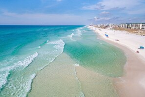 Ein Strand mit klarem türkisblauem Wasser, einem Sandufer und Gebäuden im Hintergrund.