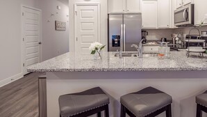 Una cocina moderna con armarios blancos, un refrigerador de acero inoxidable, un microondas y una encimera de granito. Hay tres taburetes de bar y un reloj en la pared.