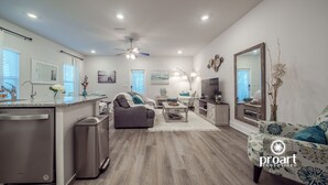 Una sala de estar moderna con un sofá gris, una mesa de centro y un televisor. Cuenta con una zona de cocina con lavavajillas y refrigerador. El ambiente tiene pisos de madera y un ventilador de techo.