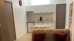 A cozy living room with a beige sofa, two red cushions, a coffee table with a glass of milk and a remote control, and a kitchen in the background with white cabinets and a microwave.