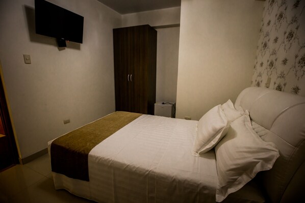Un dormitorio con cama, armario, televisor montado en la pared y una mesita de noche.