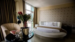 Una habitación de hotel con una cama curvada, una zona de estar con una silla y una mesa, un jarrón con flores y una ventana con cortinas.