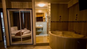 Un baño moderno con una bañera grande, una zona de ducha separada y un cristal que da a un dormitorio con cama y toalla.