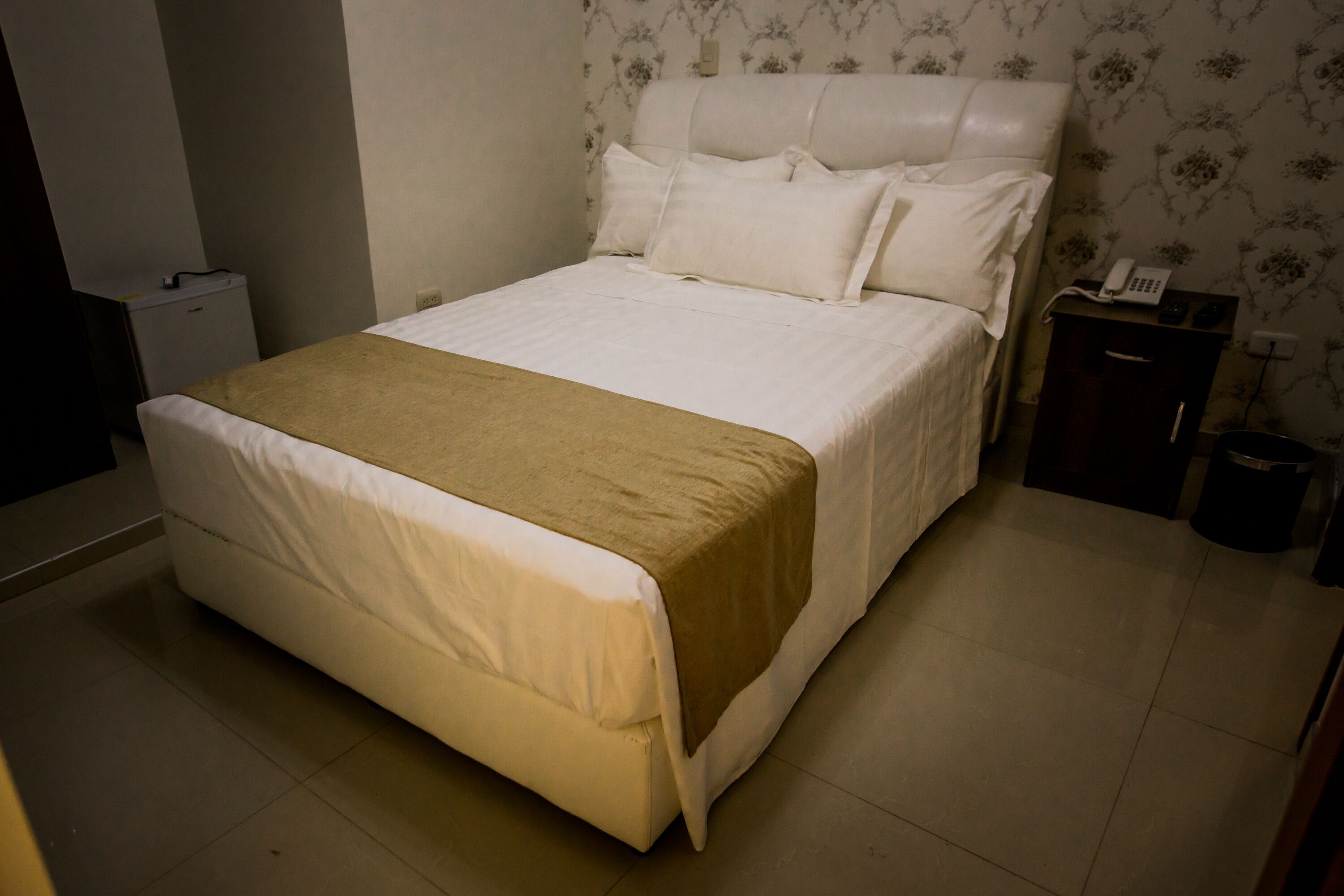 Una cama bien hecha con una manta color beige, una mesita de noche con un teléfono y un cesto de basura.