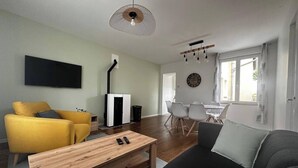A modern living room with a sofa, yellow armchair, and a wooden coffee table. A fireplace is visible in the background, and there is a dining area with a table and chairs. - Le Biardais - Gites du Bessin (Balleroy-sur-Drôme)