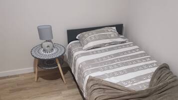 Un llit individual amb un edredó estampat, una taula de nit amb una làmpada i un tamboret estampat.