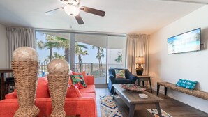 A living room with a red sofa, a coffee table, and a TV on the wall. There is a balcony with a view of palm trees. - Sandcastle Condominiums Unit 125  (Port Aransas)