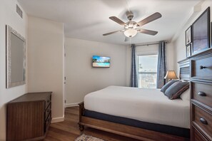 A bedroom with a bed, a ceiling fan, a TV, a dresser, and a window with curtains. - Sandcastle Condominiums Unit 125 (Port Aransas)