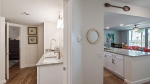 A kitchen with white cabinets, granite countertops, and a large island. There is a dining area with a red couch and a view of the outdoors. - Sandcastle Condominiums Unit 125  (Port Aransas)
