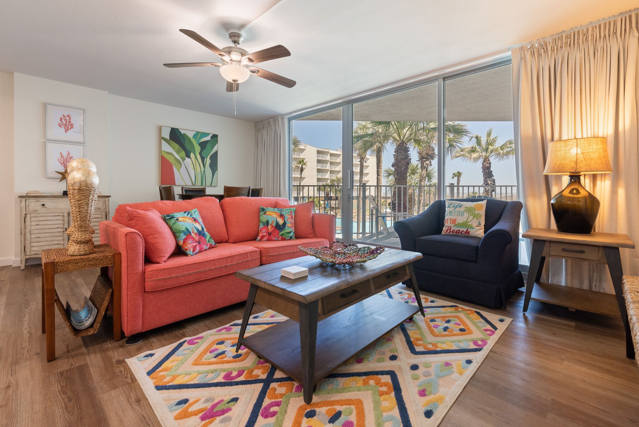 A living room with a red sofa, a blue armchair, a wooden coffee table, and a large window with a view of palm trees and buildings.