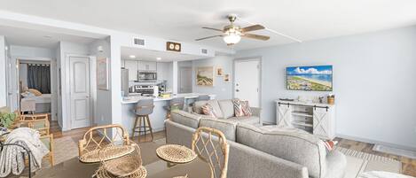 A modern living room with a grey sofa, wicker chairs, a dining table, and a kitchen in the background.