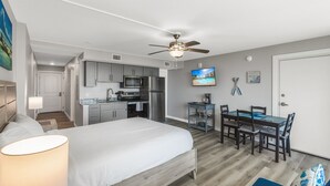 A bedroom with a bed, a bedside lamp, a ceiling fan, and a view into the adjacent kitchen area. - Sandcastle Condominiums Unit 207  (Port Aransas)