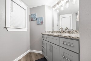 A bathroom with a granite countertop, a sink with a faucet, a mirror, and two wall-mounted light fixtures. - Sandcastle Condominiums Unit 207  (Port Aransas)