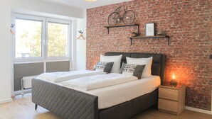 A modern bedroom with a brick wall, a large bed with white bedding, a bicycle decoration, and a bedside table with a lamp. - New York style, parking lot, lockable bike box with charging facility  (Koblenz)
