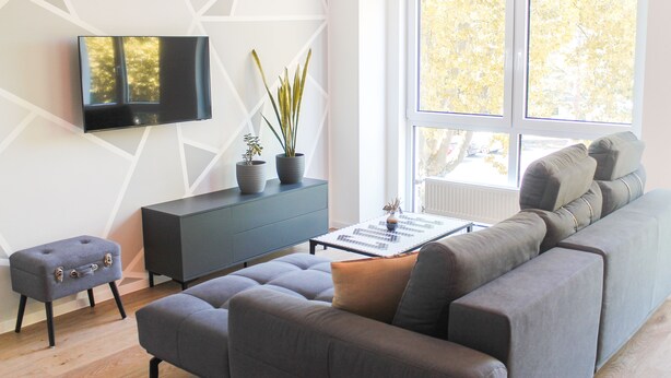 A modern living room with a grey sofa, a small ottoman, a black cabinet, a flat-screen TV mounted on the wall, and a large window with a view of trees. - New York style, parking lot, lockable bike box with charging facility  (Koblenz)