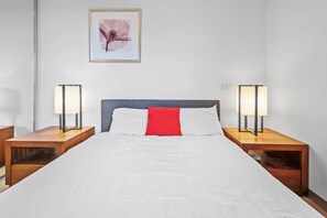 A hotel room with a bed, two bedside tables, two lamps, a red pillow, and a framed picture on the wall.