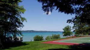 A tennis court with a red surface, surrounded by green grass and trees, overlooking a body of water and distant hills.