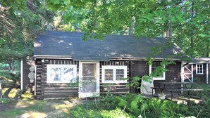 A rustic log cabin with a porch, surrounded by lush greenery and a stone pathway.