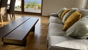 Sala de estar com mesa de centro de madeira, sofá cinza com almofadas amarelas e vista para um jardim através de uma porta deslizante.