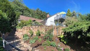 Un barrio residencial con un muro de piedra, un jardín con diversas plantas y un edificio blanco con techo verde.