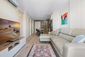 Un salon moderne doté d’un grand téléviseur à écran plat, d’un canapé beige, d’un tapis à motifs et d’un coin cuisine à l’arrière-plan.